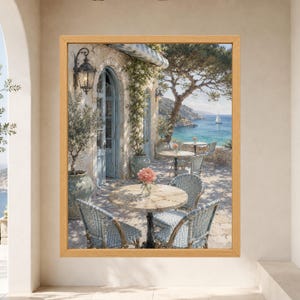 May include: A framed wall art print depicting a Mediterranean terrace with light blue wicker chairs, round stone tables, a potted plant, and a stone building with an arched doorway, overlooking a bright blue sea with a distant sailboat.