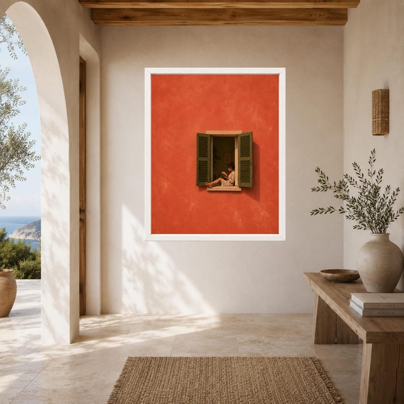 May include: A framed wall art print featuring a vibrant orange exterior wall with an open window. The window has dark green shutters and shows a person sitting inside reading a book. The artwork is displayed in a white frame on a neutral wall.