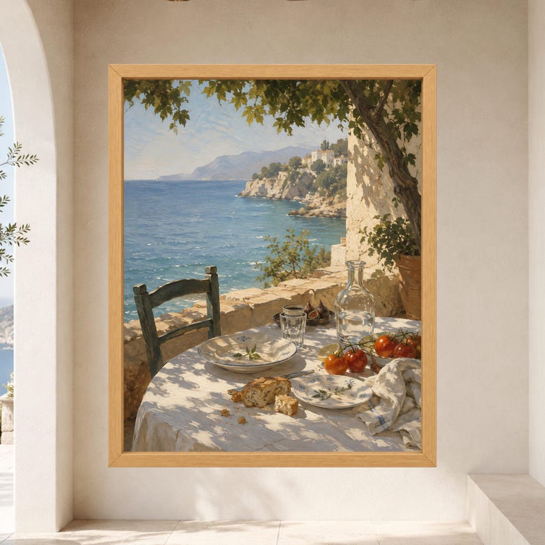 May include: A framed wall art print depicting a coastal scene with a table set for a meal. The table features a white cloth, a plate with bread, a glass carafe, and fresh tomatoes. A wooden chair sits beside the table overlooking the blue sea.