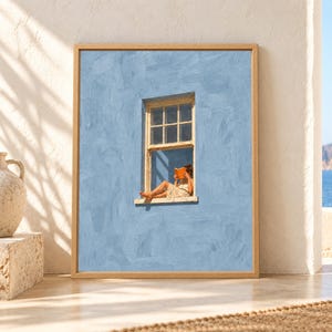 Framed soft blue wall art showing a barefoot woman sitting in an open cream window while reading an orange book against a textured minimalist blue wall.