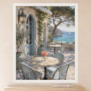 May include: A framed wall art print depicting a Mediterranean terrace with stone flooring, round marble tables, and woven blue and white chairs. A large potted olive tree, climbing vines, and a view of the blue sea with a sailboat are visible.