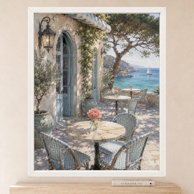 May include: A framed wall art print depicting a Mediterranean terrace with stone flooring, round marble tables, and woven blue and white chairs. A large potted olive tree, climbing vines, and a view of the blue sea with a sailboat are visible.