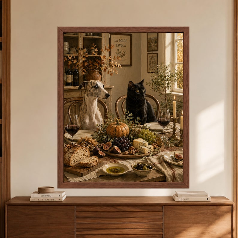Framed autumn dining wall art showing a brindle greyhound and black cat seated at a rustic harvest table with red wine, figs, grapes, cheese, bread, olives and a small pumpkin in a warm countryside interior.