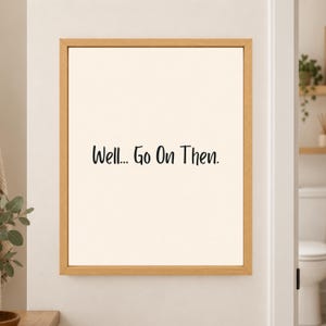 Framed minimalist bathroom wall art with the handwritten black phrase “Well... Go On Then.” centered on a warm off-white background in a light wood frame beside a bathroom doorway.