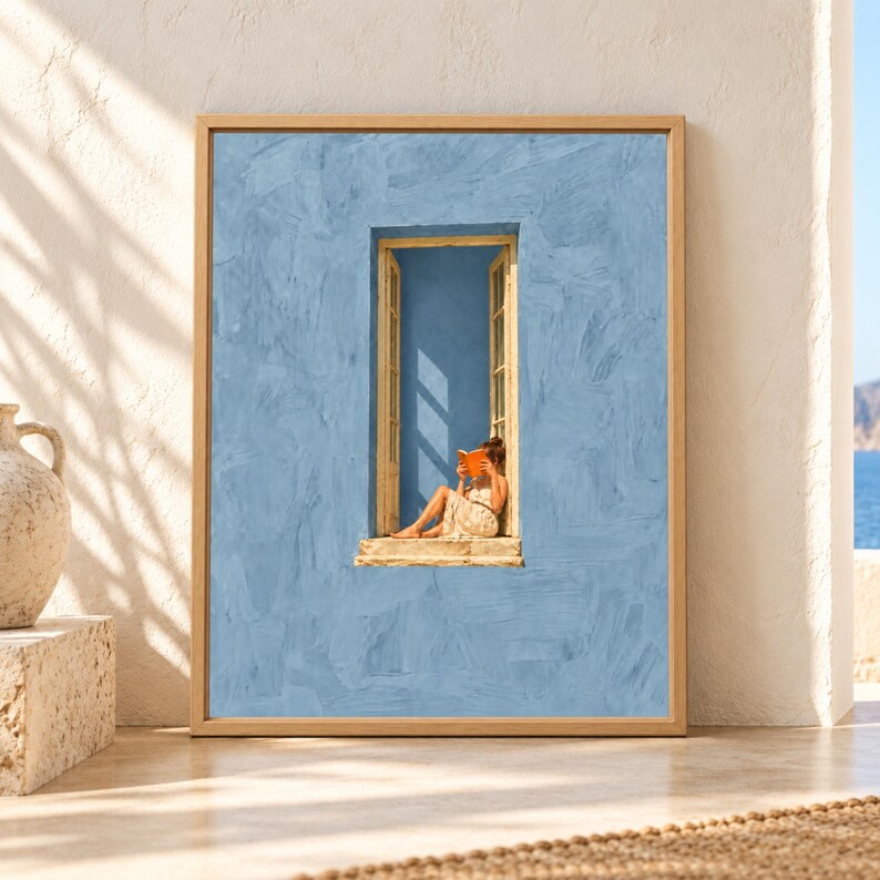 Framed blue wall art showing a barefoot woman reading an orange book in a tall narrow open window surrounded by a large textured soft blue Mediterranean facade.