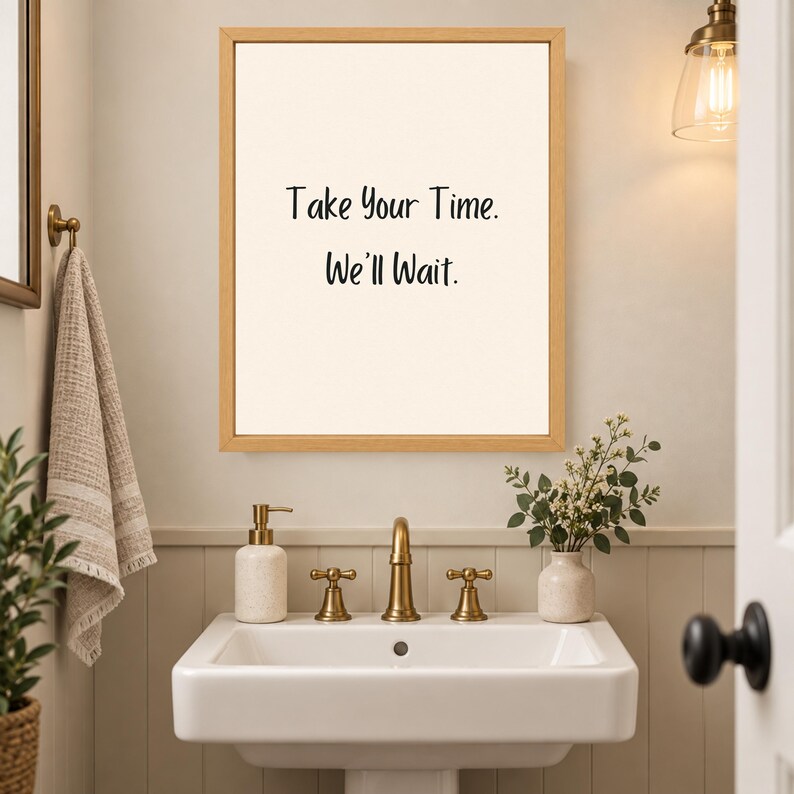 May include: A rectangular wall art piece in a light wood frame featuring a cream-colored background with the black text Take Your Time. We'll Wait. centered on the print. The artwork is displayed above a white bathroom sink with gold fixtures.
