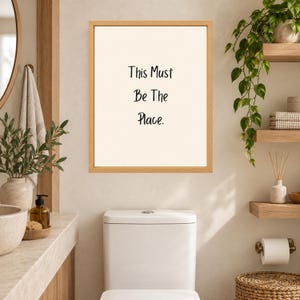 May include: A rectangular wall art piece in a light wood frame featuring a cream-colored background with the black text This Must Be The Place centered in the middle. The artwork is displayed on a wall above a white toilet in a bathroom setting.