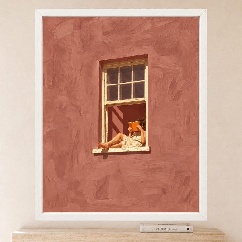May include: A framed wall art print featuring a textured, terracotta-colored wall with a single window. A person sits in the window frame, wearing a light-colored dress and reading an orange book. The artwork is enclosed in a simple white frame.