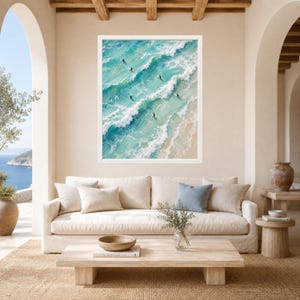 May include: A framed wall art print featuring an aerial view of turquoise ocean waves crashing onto a sandy beach. Several surfers are visible in the water, riding the waves. The artwork is displayed in a white frame on a beige wall.