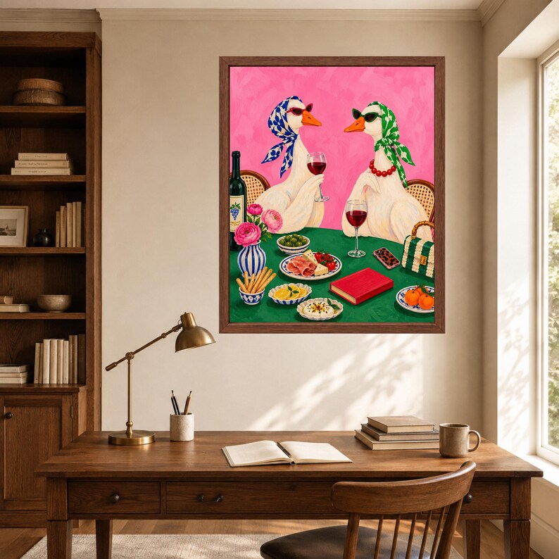 May include: A framed wall art print featuring two white geese wearing headscarves and sunglasses, sitting at a green table with wine, snacks, and a red book. The background is a vibrant pink, and the geese are depicted in a whimsical, colourful style.