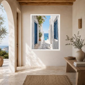 K&ouml;nnte beinhalten: Ein gerahmter Kunstdruck, der eine schmale mediterrane Gasse mit wei&szlig;en Stuckw&auml;nden, leuchtend blauen T&uuml;ren und Fensterl&auml;den, einer Topfpflanze und einem Steinweg zeigt, der unter klarem Himmel zu einem hellblauen Meer f&uuml;hrt.