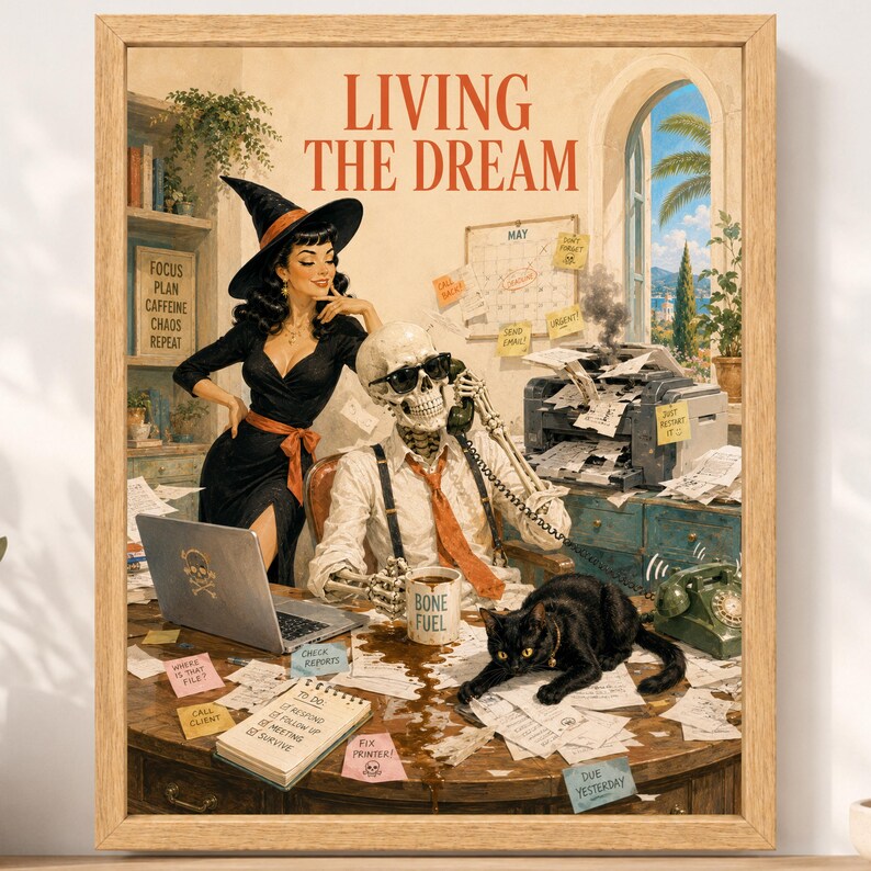 May include: A framed wall art print titled LIVING THE DREAM featuring a person in a black dress and witch hat standing behind a skeleton wearing sunglasses and a white shirt. A black cat sits on a desk covered in papers, a laptop, and a phone.