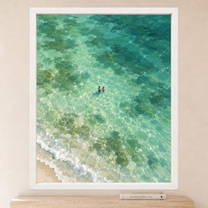 May include: A framed wall art print featuring an aerial view of a sandy beach shoreline with turquoise and emerald ocean water. Two small figures are swimming in the clear, shallow sea, with visible light reflections on the sandy ocean floor.
