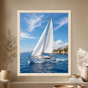 Framed Mediterranean sailboat wall art showing a modern white sailing yacht with full sails crossing deep blue water beside a rocky coastline with white hillside houses under a clear blue sky.