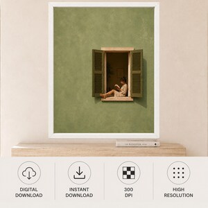 May include: A framed wall art print featuring a sage green textured wall with an open window. The window has dark green shutters and shows a person sitting on the ledge reading a book. The frame is white and the print is displayed on a wooden shelf.