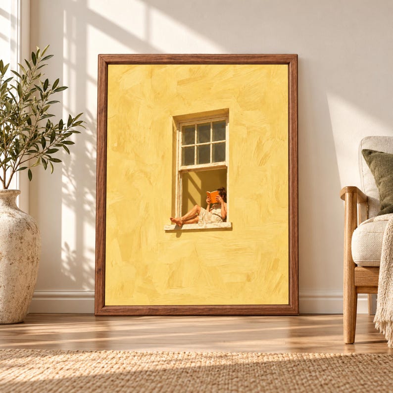 Framed yellow wall art showing a barefoot woman sitting in a small open window reading an orange book against a large textured sunlit yellow facade.