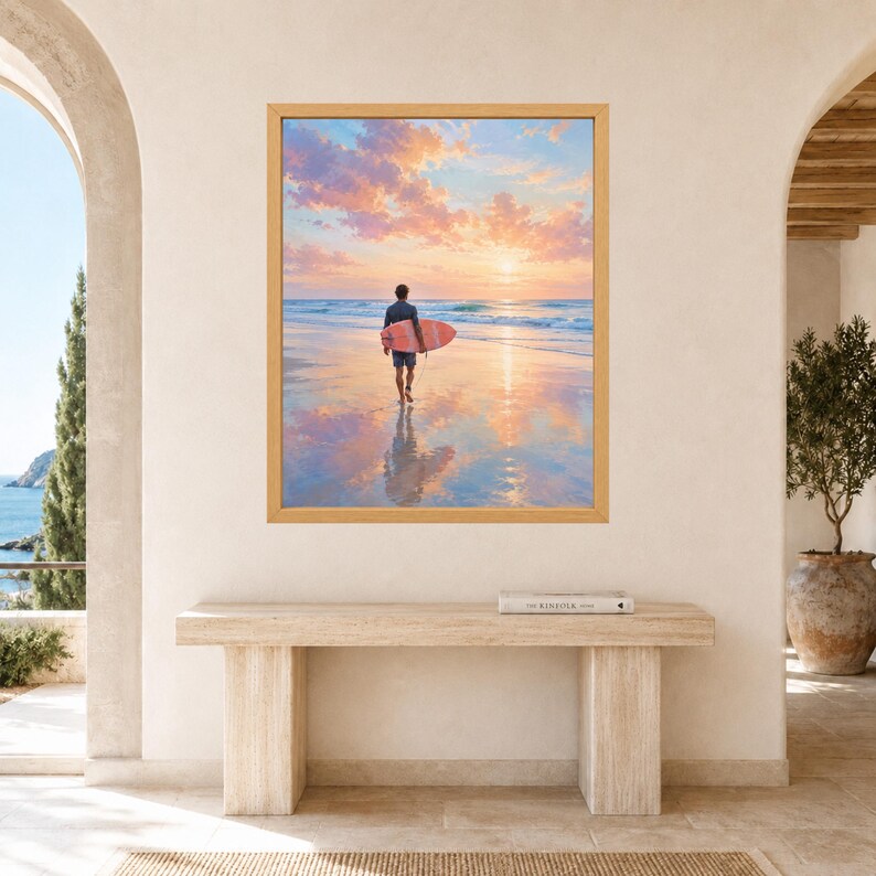 May include: A framed wall art print depicting a person walking on a sandy beach at sunset while carrying a pink surfboard. The scene features a calm ocean, soft clouds in shades of pink and orange, and a clear reflection on the wet sand.