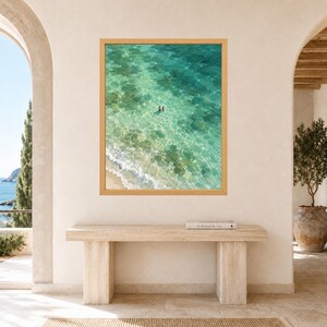 May include: A framed wall art print featuring an aerial view of a turquoise ocean with clear water, sandy shoreline, and two small figures swimming in the center. The artwork is encased in a light-toned wooden frame and displayed on a wall.