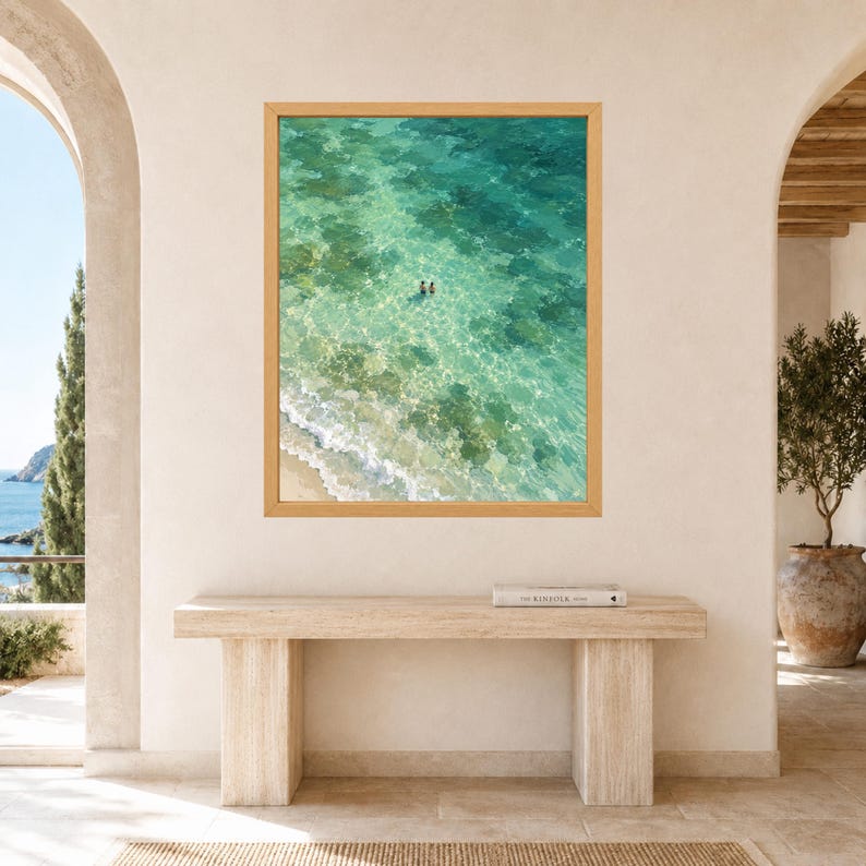 May include: A framed wall art print featuring an aerial view of a turquoise ocean with clear water, sandy shoreline, and two small figures swimming in the center. The artwork is encased in a light-toned wooden frame and displayed on a wall.