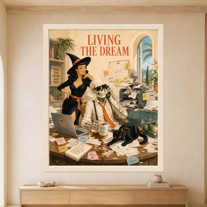 Framed Living the Dream wall art showing a skeleton on the phone at a chaotic office desk with spilled coffee, smoking printer, paperwork, a retro witch and a black cat lying across the work.