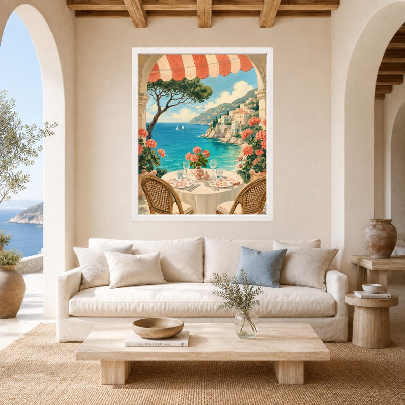 May include: A framed wall art print depicting a coastal Mediterranean scene with a round table set for two under a red and white striped awning, overlooking a blue sea with distant white buildings, green trees, and pink flowers in the foreground.