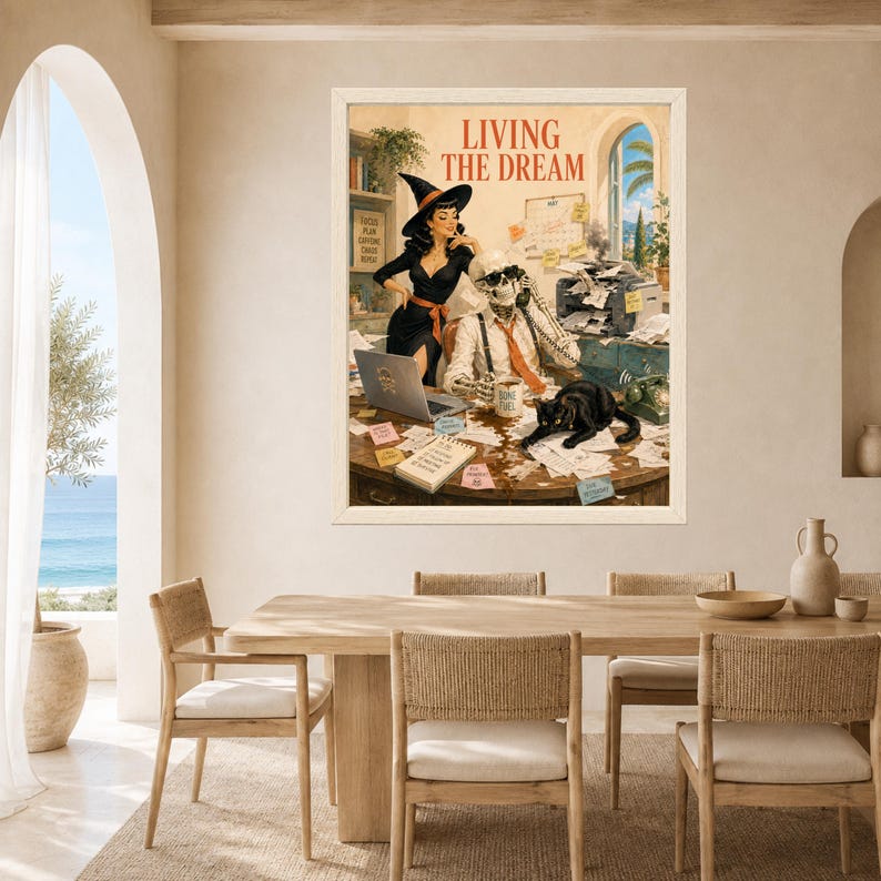 May include: A framed wall art print titled LIVING THE DREAM featuring a witch in a black dress and a skeleton in a white shirt and orange tie sitting at a cluttered desk with a laptop, a black cat, and office supplies in a warm-toned room.