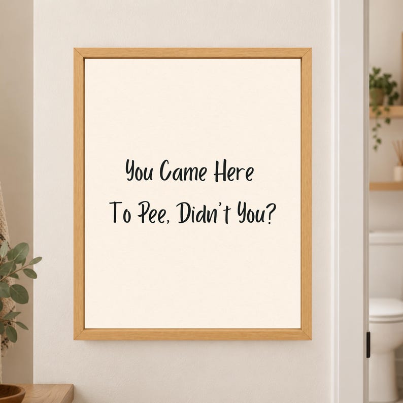 Framed minimalist bathroom wall art with the handwritten black phrase “You Came Here To Pee, Didn’t You?” centered on a warm off-white background in a light wood frame beside a bathroom doorway.