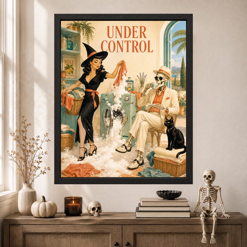 May include: A framed wall art print titled UNDER CONTROL featuring a witch in a black dress and hat doing laundry with a skeleton in a beige suit and hat sitting nearby drinking tea. A black cat sits on a wicker basket filled with folded towels.