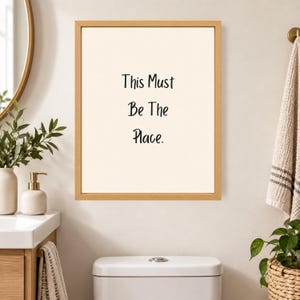 May include: A rectangular wall art piece featuring a light wood frame and a cream-colored background. The text "This Must Be The Place." is printed in black, sans-serif lettering in the center of the print, creating a clear and readable display.