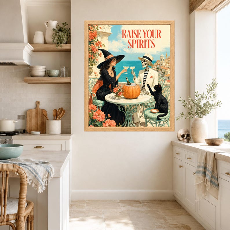 May include: A framed wall art print featuring a witch in a black dress and a skeleton in a white suit sitting at a table with a pumpkin and a black cat. The text RAISE YOUR SPIRITS is printed at the top. The scene is set on a balcony overlooking the sea.