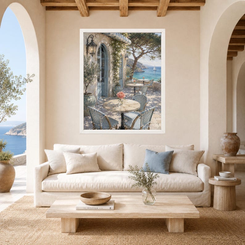 May include: A framed wall art print depicting a Mediterranean coastal terrace with light blue wicker chairs, round wooden tables, a stone archway, and a view of the sea with a sailboat under a clear sky, displayed above a beige sofa in a room.
