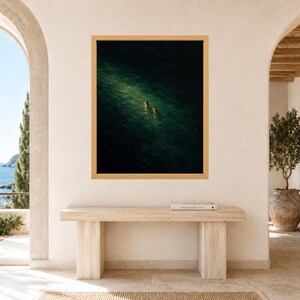 May include: A framed wall art print featuring an aerial view of dark green ocean water with two small figures swimming in a patch of bright sunlight. The artwork is displayed in a light wood frame hanging on a plain, light-colored interior wall.