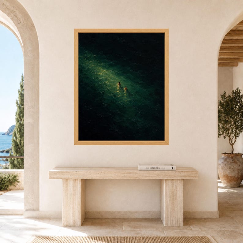 May include: A framed wall art print featuring an aerial view of dark green ocean water with two small figures swimming in a patch of bright sunlight. The artwork is displayed in a light wood frame hanging on a plain, light-colored interior wall.
