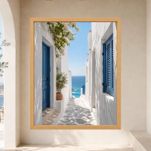 Framed Greek island coastal wall art showing a sunlit whitewashed stone alley with cobalt blue doors and shutters leading toward the deep blue Mediterranean sea beneath flowering bougainvillea.