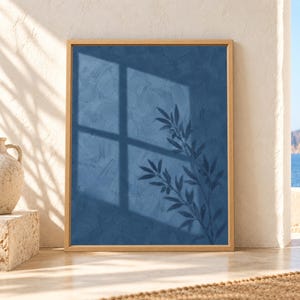 Framed blue botanical wall art showing an olive branch silhouette and large window shadow across a textured deep blue wall in soft natural sunlight.