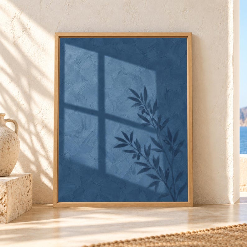 Framed blue botanical wall art showing an olive branch silhouette and large window shadow across a textured deep blue wall in soft natural sunlight.