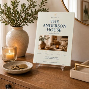 May include: A cream-coloured welcome sign for The Anderson House stay guide, featuring dark blue text, a central photograph of a living room, and five icons. The sign is displayed on a clear acrylic stand, accompanied by a small dish with metal keys.