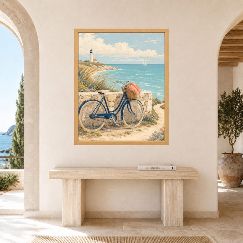 Op de afbeelding: Een ingelijste kunstdruk met een blauwe fiets met een rieten mand geparkeerd op een stenen pad bij een kustklif. Op de achtergrond staat een witte vuurtoren op een met gras begroeide heuvel die uitkijkt over de blauwe oceaan met een zeilboot.