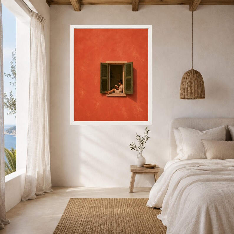 May include: A framed wall art print featuring a vibrant orange wall with an open window. The window has dark green shutters and shows a person sitting on the ledge reading a book. The artwork is displayed in a white frame on a neutral wall.