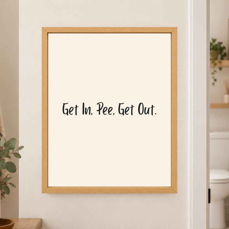 Framed minimalist bathroom wall art with the handwritten black phrase “Get In, Pee, Get Out.” centered on a warm off-white background in a light wood frame beside a bathroom doorway.