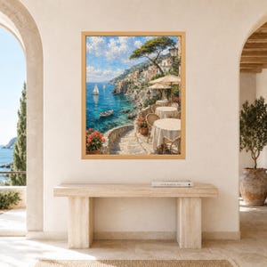 Pu&ograve; includere: Una stampa artistica da parete incorniciata che raffigura una scena costiera mediterranea con una terrazza in pietra, tavoli rotondi con tovaglie beige e sedie in vimini che si affacciano su un mare blu con barche a vela e un villaggio.