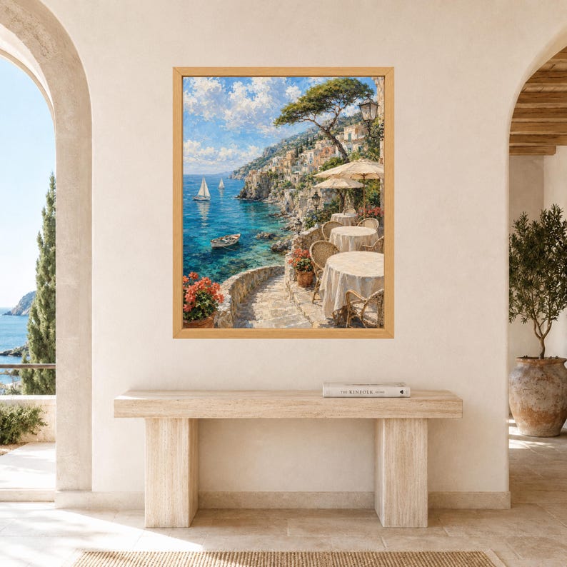 Pu&ograve; includere: Una stampa artistica da parete incorniciata che raffigura una scena costiera mediterranea con una terrazza in pietra, tavoli rotondi con tovaglie beige e sedie in vimini che si affacciano su un mare blu con barche a vela e un villaggio.