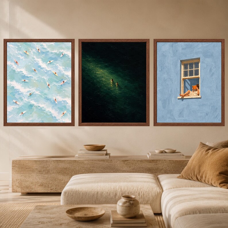 May include: A set of three square framed wall art prints. The left print shows surfers on blue ocean waves. The center print depicts two figures swimming in dark green water. The right print features a person reading in a window on a blue wall.
