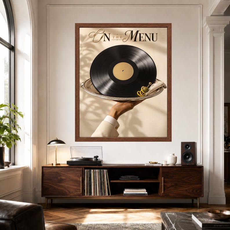 May include: A framed wall art print titled On The Menu featuring a black vinyl record served on a silver platter. The record has a gold center label and is garnished with a yellow lemon twist. The background is a light cream color with shadows.