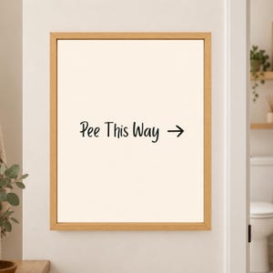 Framed minimalist bathroom wall art with the handwritten black phrase “Pee This Way” and a right-facing arrow centered on a warm off-white background in a light wood frame beside a bathroom doorway.