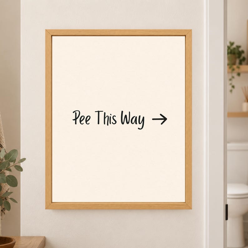 Framed minimalist bathroom wall art with the handwritten black phrase “Pee This Way” and a right-facing arrow centered on a warm off-white background in a light wood frame beside a bathroom doorway.
