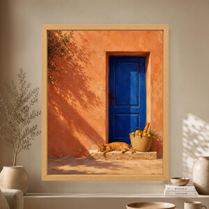 Framed Mediterranean wall art showing an orange tabby cat sleeping beside a woven market basket with bread and lemons in front of a cobalt blue door and sunlit terracotta wall.