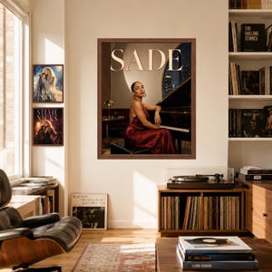 May include: A framed wall art print featuring a portrait of Sade in a red satin dress sitting at a grand piano, with the text SADE in large white letters at the top. The print is displayed on a wall alongside two smaller Stevie Nicks posters.
