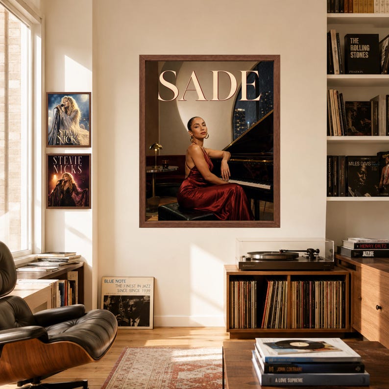 May include: A framed wall art print featuring a portrait of Sade in a red satin dress sitting at a grand piano, with the text SADE in large white letters at the top. The print is displayed on a wall alongside two smaller Stevie Nicks posters.