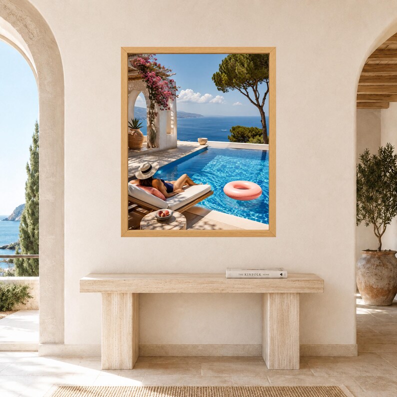 May include: A framed wall art print depicting a Mediterranean coastal scene with a person in a sun hat lounging on a white chaise longue by a blue swimming pool. A pink inflatable ring floats in the water, with bougainvillea and sea views.